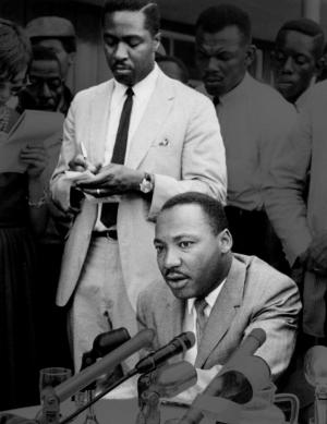 Black and white photo of Martin Luther King Jr sitting and speaking with Dr. Clarence B. Jones standing behind him.