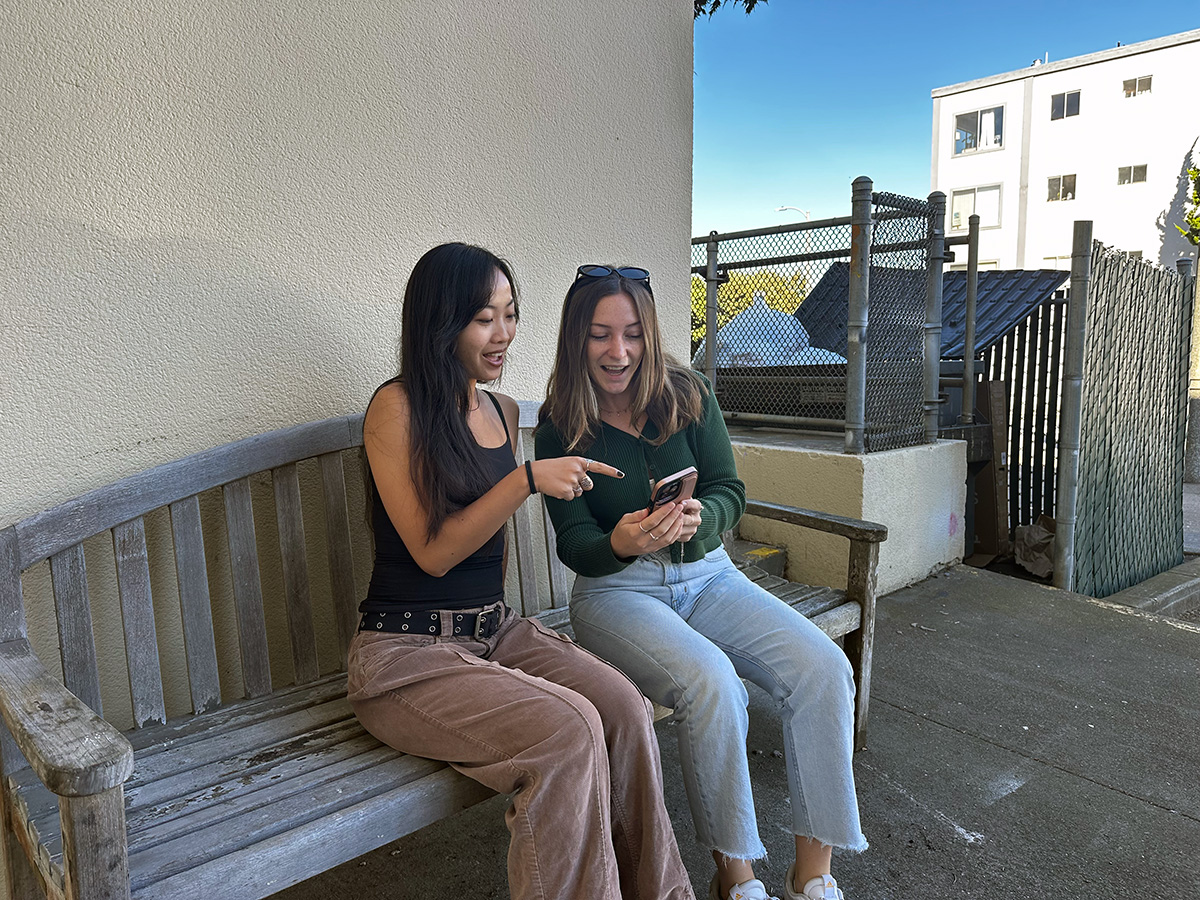 "Two students sitting on a bench looking at each others phones"