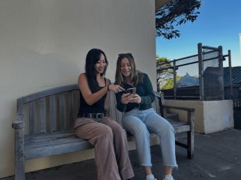 "Two students sitting at a bench outside looking at a phone"
