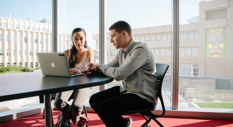 Two individuals sitting next to each, while looking over the same laptop.