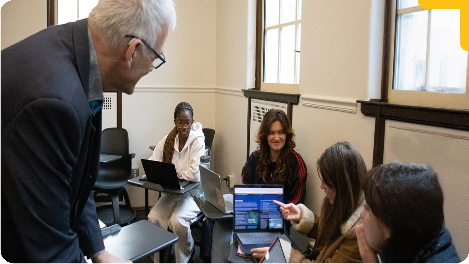 Professor Zarobell leans over to speak with a small group of students seated in a classroom. One student holds a tablet while others sit with laptops, and large windows line the room, suggesting a collaborative discussion or mentoring session.