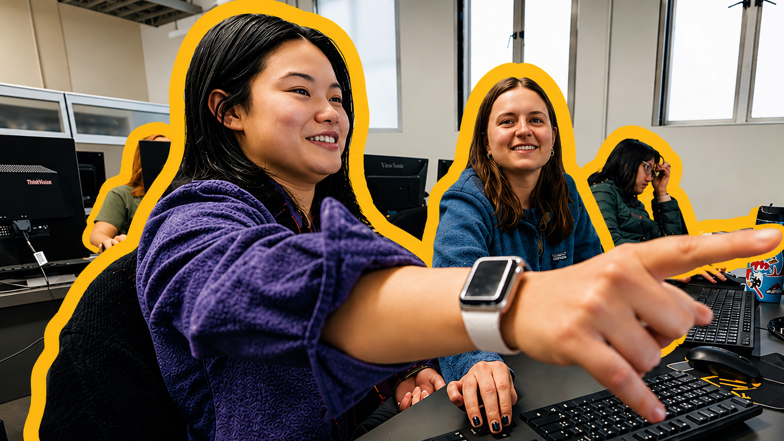Young adults seated at desktop computers in a classroom or lab setting, collaborating on work. A student in the foreground wearing a purple fleece and a smartwatch points toward something off-screen while smiling, as two other students behind her look on. The group appears engaged and focused, with monitors and keyboards visible on the desks.