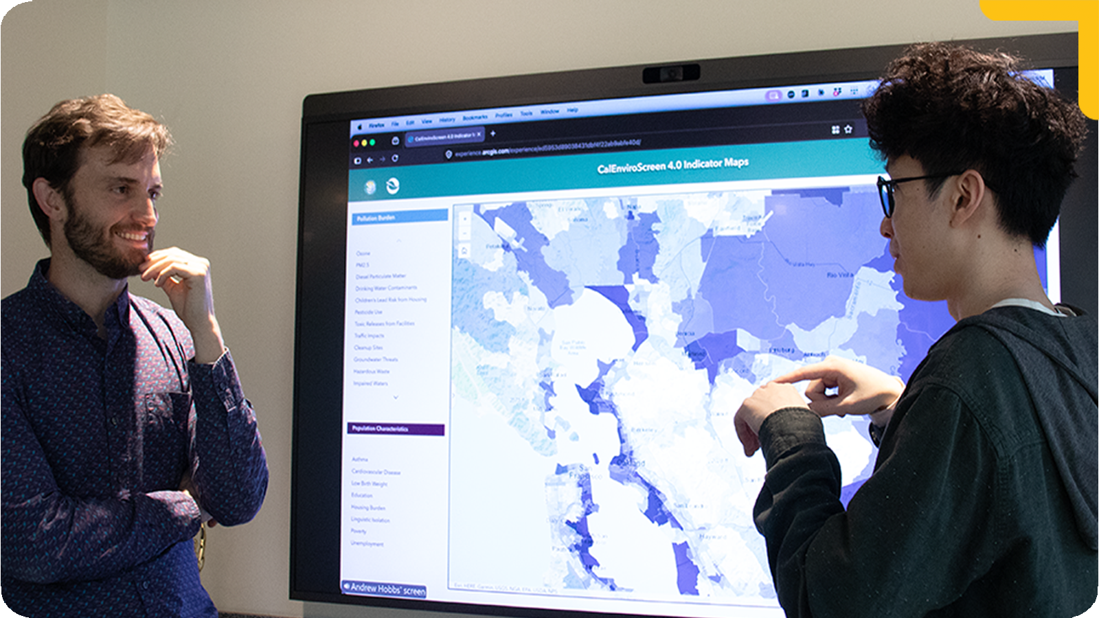 Professor Hobbs and his graduate student standing in front of a large wall-mounted display showing a detailed geographic map with shaded regions. One gestures toward the screen while explaining, and the other listens with a hand near his face. The setting appears to be a classroom or lab focused on data analysis.