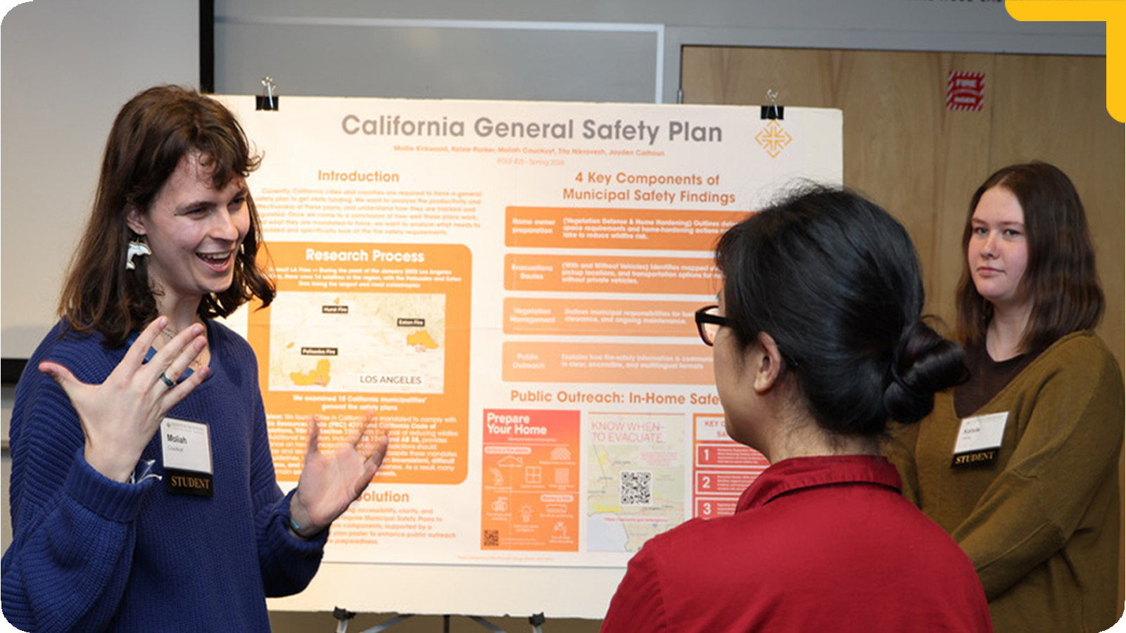 A small group of people stands in front of a large poster presentation board. One person gestures while explaining the content, while others face them and listen, indicating a discussion or critique session in an academic setting.