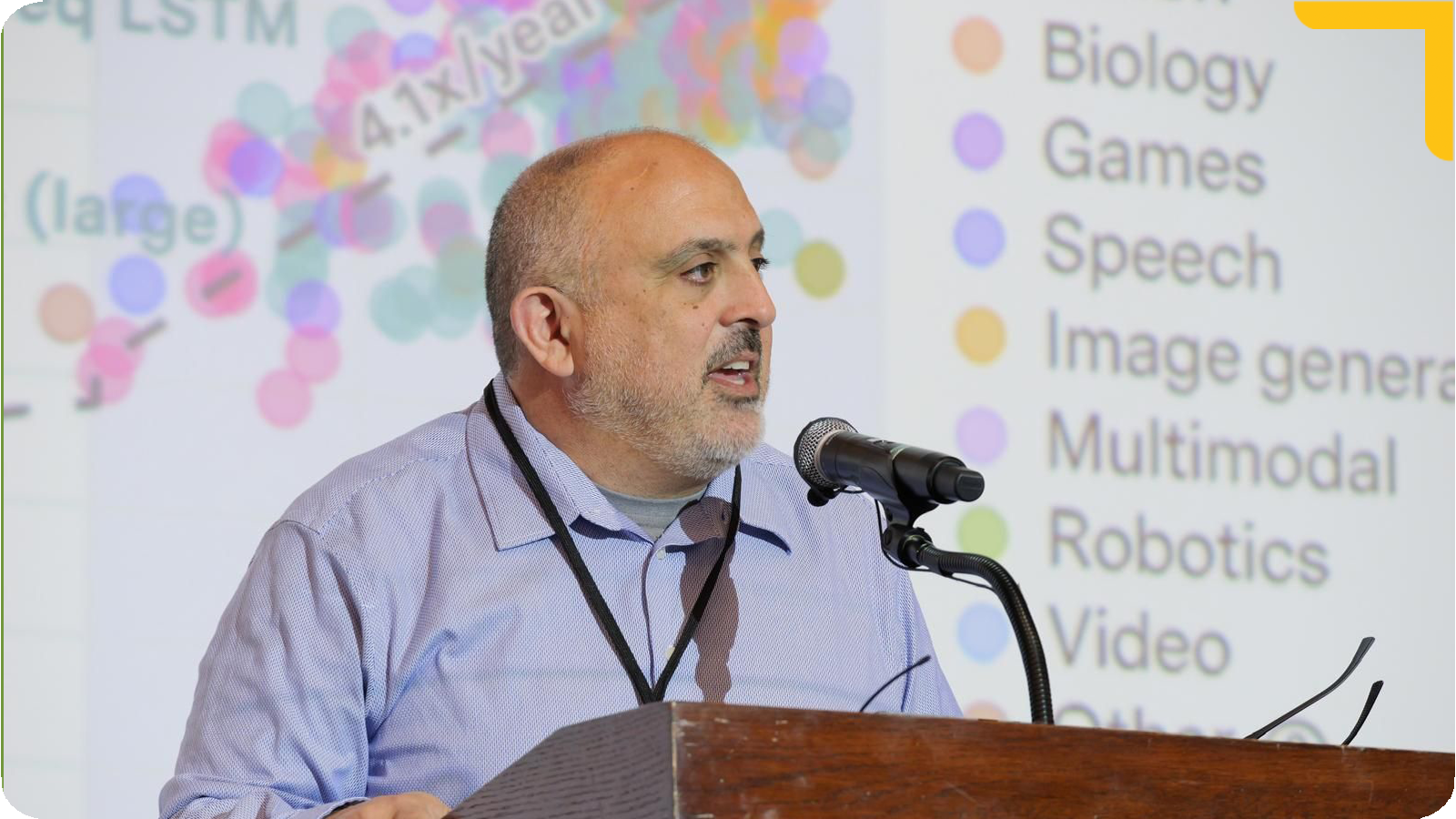 Professor Saah stands at a podium speaking into a microphone, gesturing with one hand. Behind him is a projected slide filled with colorful text and categories, suggesting a presentation on technology or research topics.
