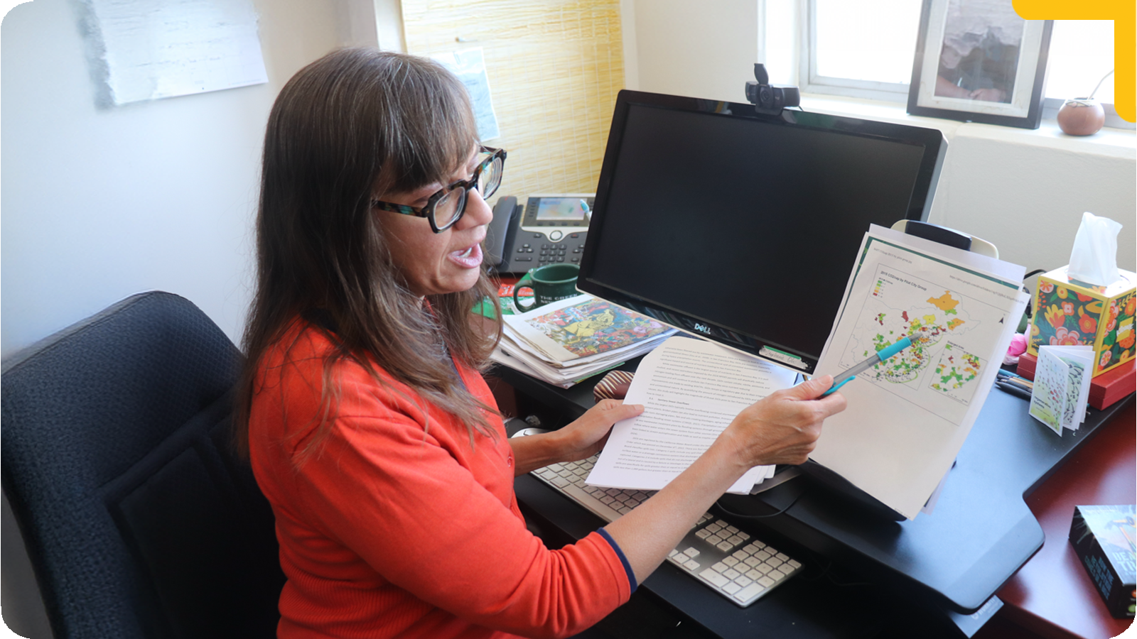 Professor Siehr sits at a desk in an office, holding and reviewing a printed sheet with a map or data visualization. A computer monitor and office items are visible nearby, suggesting focused individual work on research or policy materials.