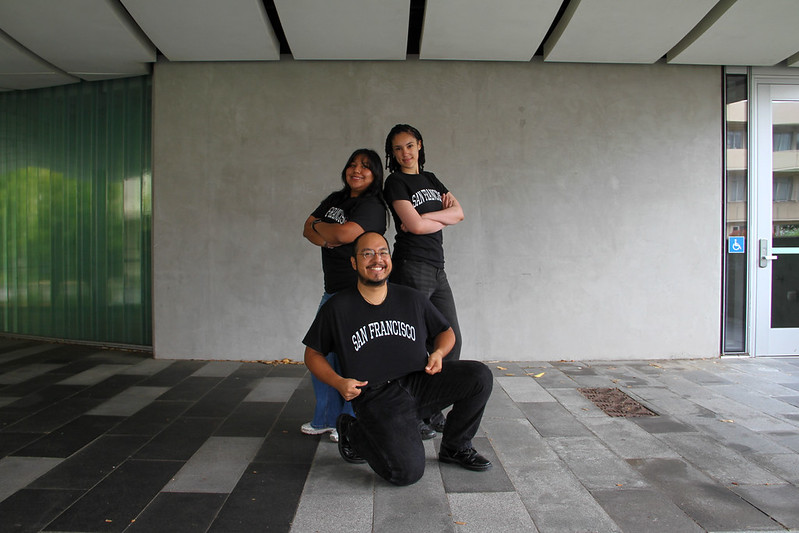 Two individuals standing back to back with arms folded, one kneeling on floor in front