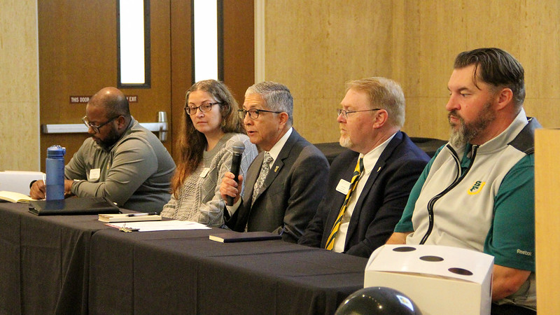 Various campus staff leaders sitting at a table with President Aceves holding a microphone answering a question.