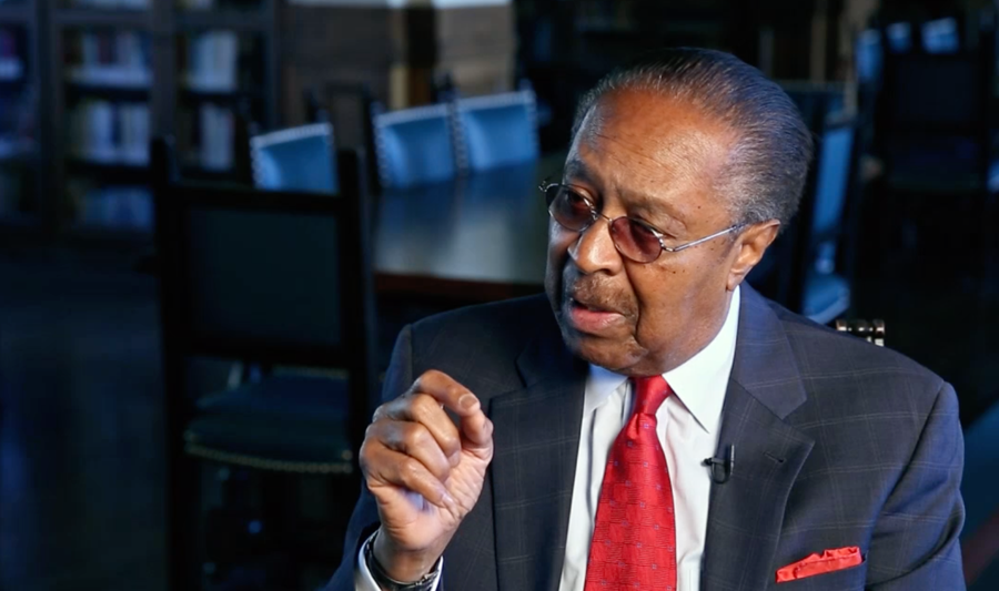 A man in a suit talks, seated in a library