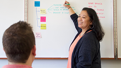 Woman smiles and points to text on a whiteboard while someone looks on