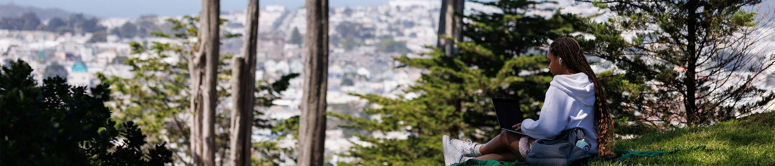 "young person sitting on a hill looking out at the city"