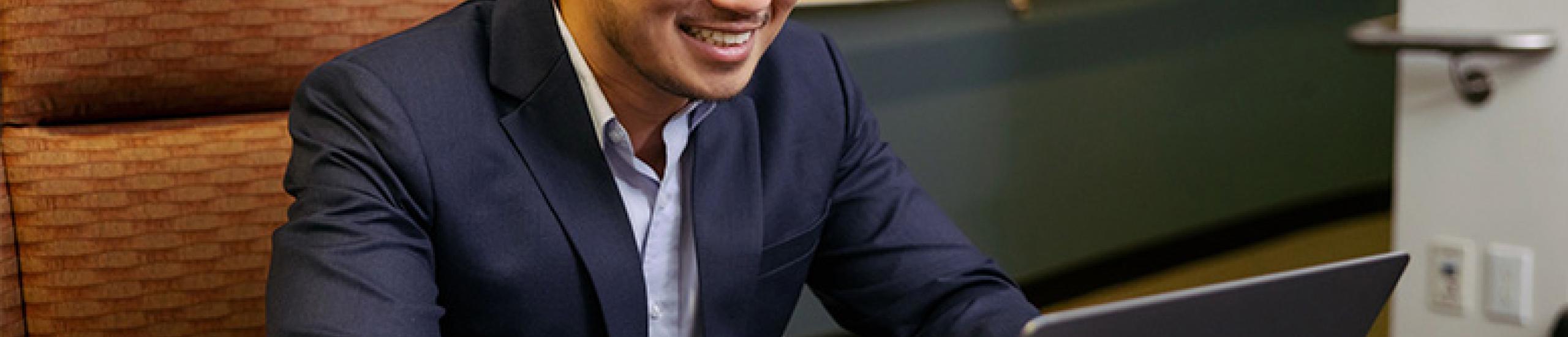 A person with short black hair is smiling while looking at their laptop. They are wearing a blue blazer and collared shirt and are sitting at a table.