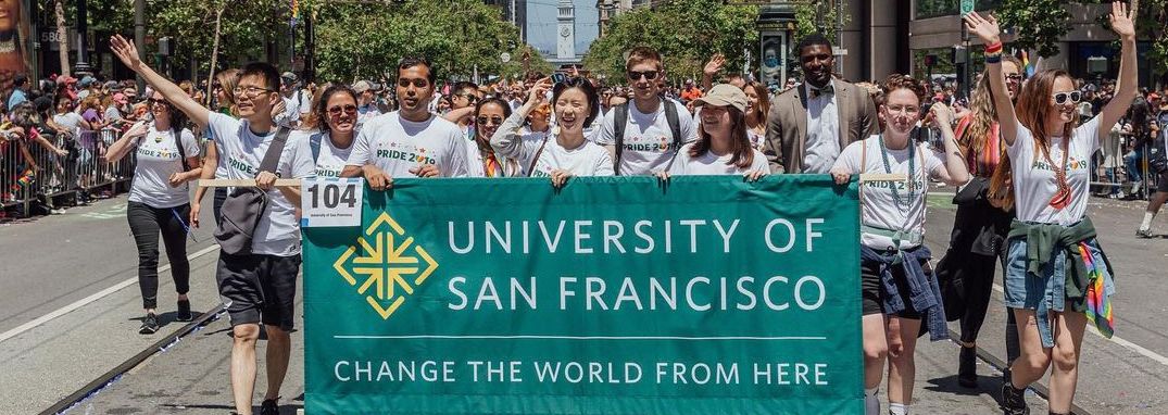 Five smiling people hold a banner that reads "University of San Francisco, Change the World From Here". Behind them is a large crowd of people who are all walking in the San Francisco Pride Parade, with the San Francisco Ferry Building Clocktower visible in the background.