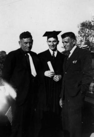 Black & white photograph of three men wearing suits, man in the middle wears a mortarboard and holds a diploma in his hand 