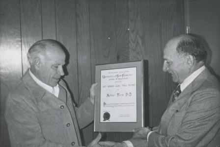 Black and white photo of two individuals shaking hands and holding a framed certificate