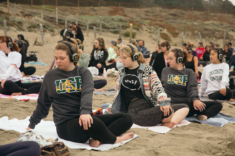 USF students sitting on the sand at Baker Beach practicing yoga with “silent disco” headphones on. The photo captures 4 USF female students in the front row, two with eyes closed, two with eyes open, all four with their left hand on the right knee, right arm placed behind them to the side, stretching. Behind them are more students immersed in the outdoor beach yoga class together.