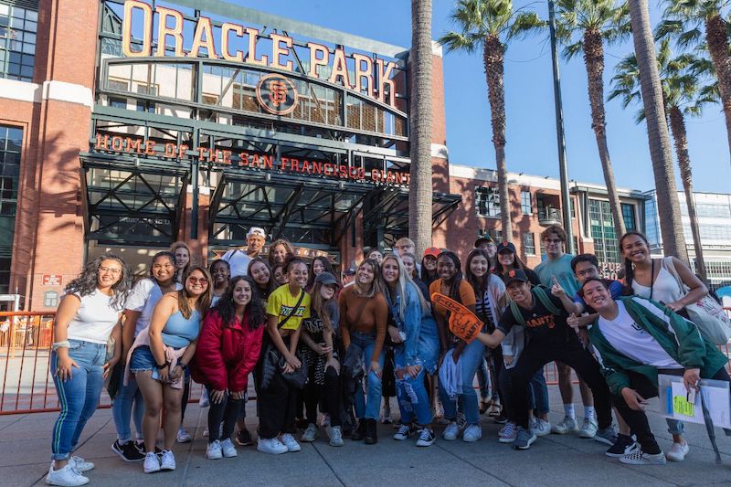 A very happy and excited group of 28 USF students standing outside of Oracle Park with palm trees behind them and the Oracle Park sign.