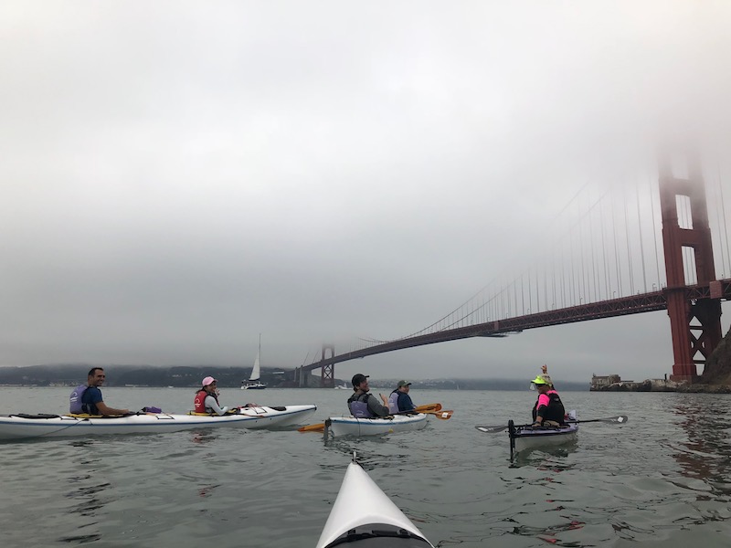 USF students in tandem kayaks headed towards the Golden Gate Bridge that’s covered in fog.