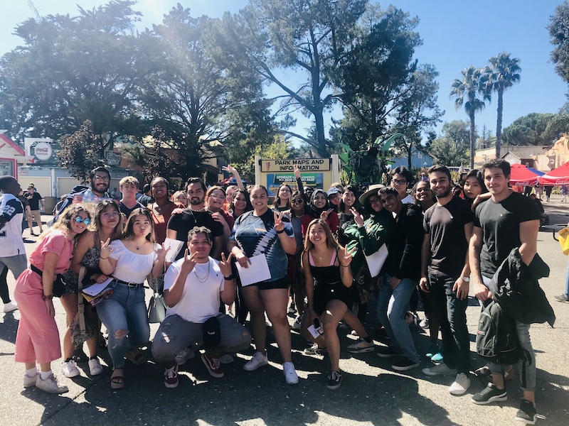 A fun and excited group of 28 USF students at Six Flags Amusement Park on a hot sunny weekend standing for a group photo outside the directory sign with trees in the background and Coca Cola sun umbrellas. 