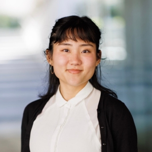"A femme Asian person smiling with their teeth closed while directly looking at the camera, they are wearing a white buttoned up blouse and a black cardigan over it. they have dark brown hair and bangs"