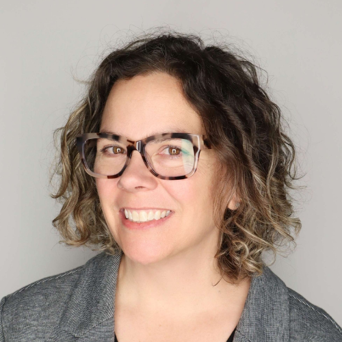 "A light-skinned woman in a grey blazer and thick framed glasses is is smiling with teeth. She has chin length curly medium brown hair."