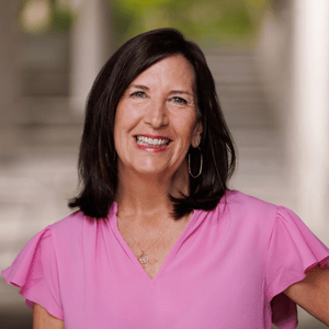"a caucasian woman with brown hair is smiling with her teeth. she is wearing a pink blouse and a gold necklace. she stands in front of a blurry background and is the point of focus in this picture"