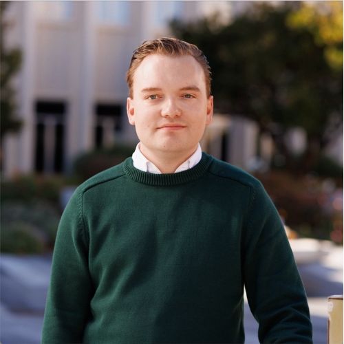 "A caucasian person in a forest green sweater is smiling with their mouth closed and looking at the camera. they have light brown and short hair swept back and are in a courtyard with green trees in the background."