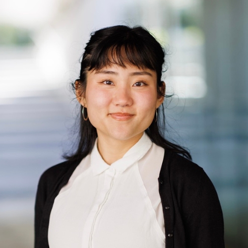 ""A femme Asian person smiling with their teeth closed while directly looking at the camera, they are wearing a white buttoned up blouse and a black cardigan over it. they have dark brown hair and bangs""