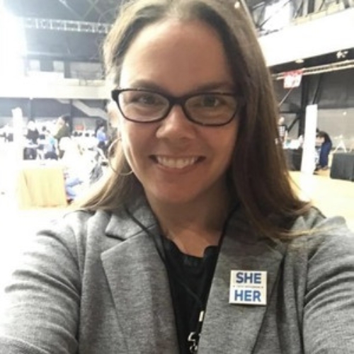 ""A light-skinned woman in a grey blazer and black framed glasses is holding her arms together to take a selfie. She is also wearing a badge that says 'she' and 'her' over each other. She has medium length straight medium brown hair. She is wearing silver hoop earrings and has a black lanyard on her neck."