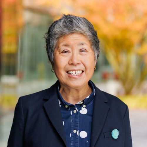 "asian woman with black blazer, short gray and black hair, smiling, blue shirt with white polka dots, blurry background of orange leaves on tress in the fall"