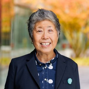 "asian woman with black blazer, short gray and black hair, smiling, blue shirt with white polka dots, blurry background of orange leaves on tress in the fall"