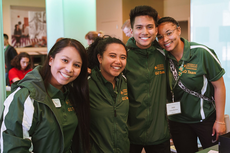 Four students smiling at the camera wearing green jackets.