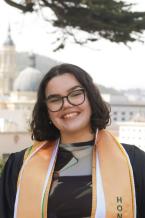 Photo of woman with short dark hair and glasses wearing a graduation gown and gold stole
