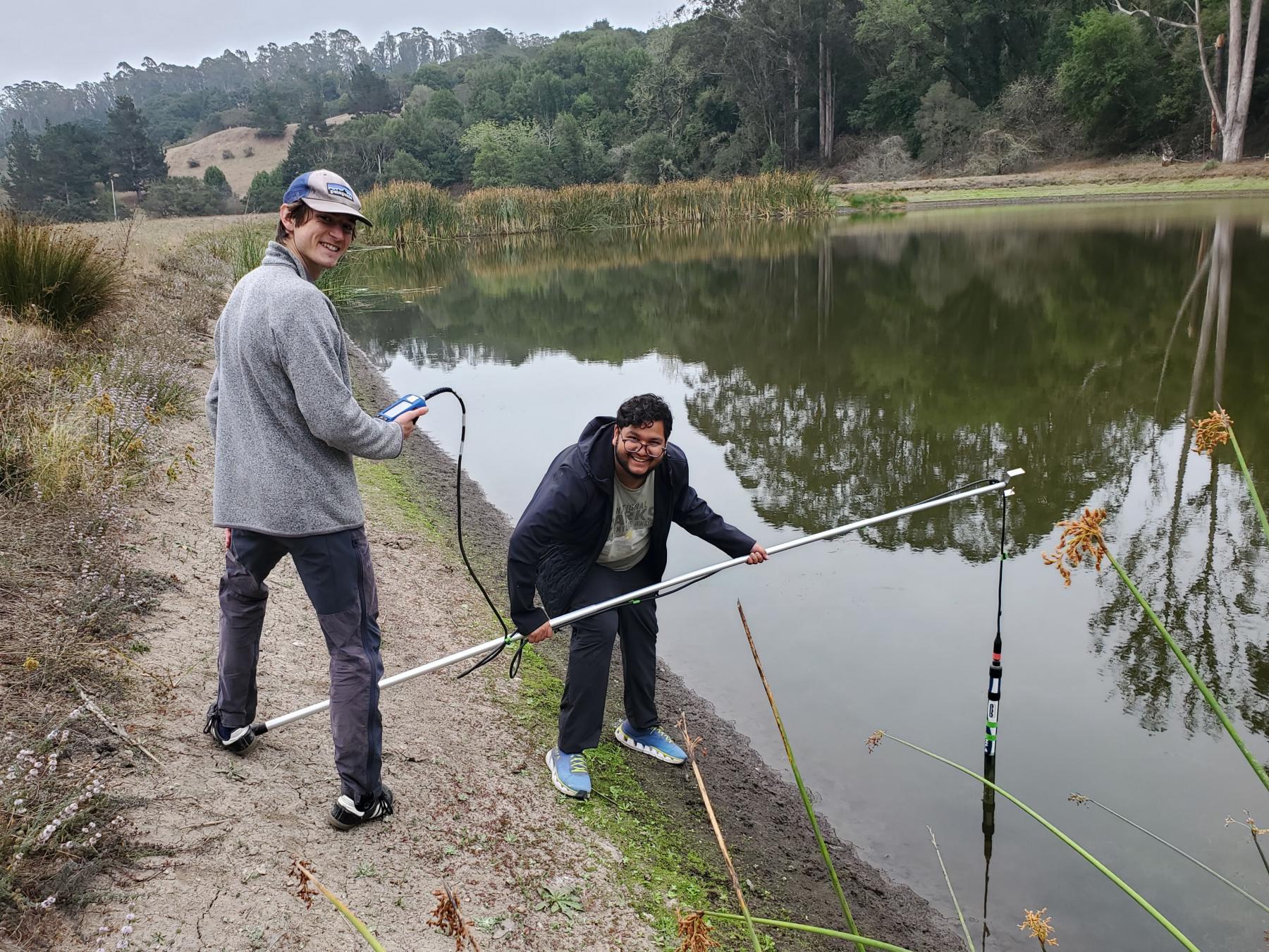 Two Environmental Science students smiling for a picture while conducting field tests by a lake. They are holding a long rod with a measuring instrument attached at the end.