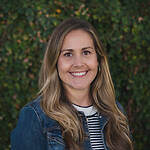 Jessica Kench is wearing a black and white striped sweater and a denim jacket. She is smiling in front of a leafy brick wall