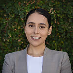 Susana Pineda is wearing a neutral beige blazer over a white shirt and is smiling in front of a leafy brick wall.